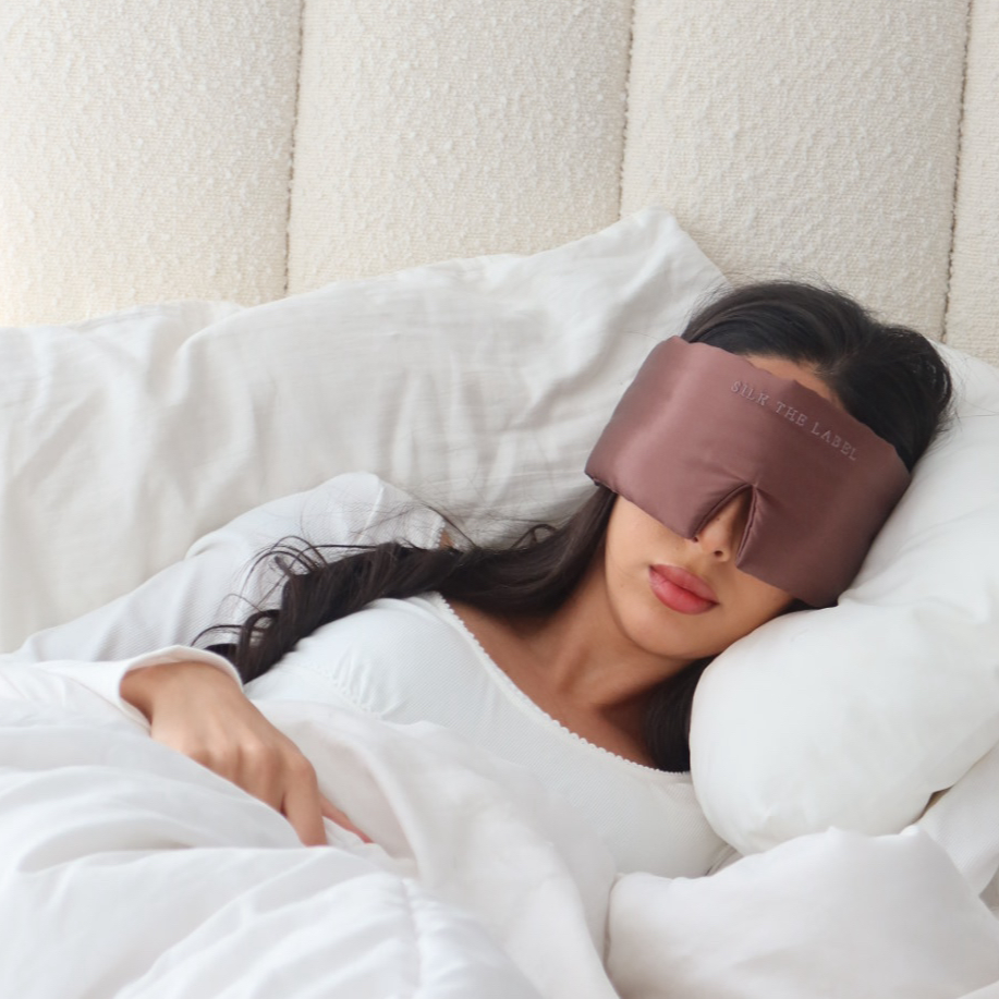 Woman lying in bed with a sleep mask on, wrapped in white bedding.